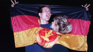 Ein jubelndes Paar hat sich 1990  am Tag der Deutschen Einheit in einer DDR-Fahne dort Platz geschaffen, wo früher das Emblem Hammer und Sichel war –  und feierte frenetisch den historischen Anlass. Foto: dpa/Zentralbild
