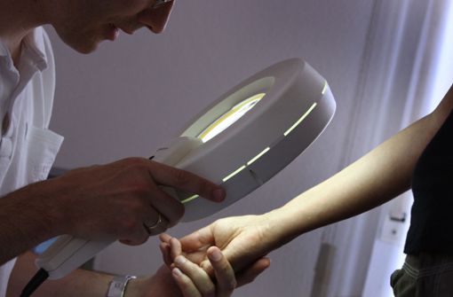 Ein Hautarzt untersucht in seiner Praxis mit einem Vergrößerungsglas die Haut einer Patientin bei einer Hautkrebs-Früherkennung. Foto: picture alliance/dpa