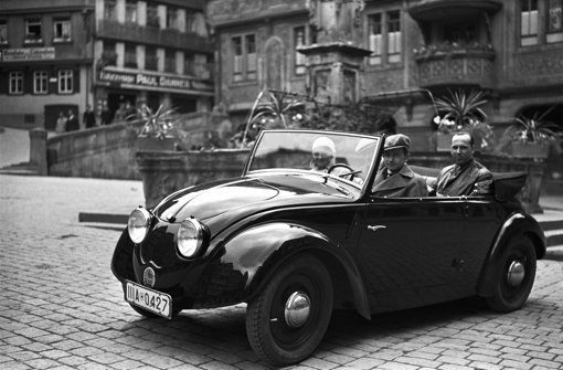 Das Werksfoto aus dem Jahr 1936 zeigt Ferry Porsche am Steuer des zweiten Volkswagen-Prototypen (V2), fotografiert auf dem Marktplatz in Tübingen. Neben ihm seine Frau Dorothea. Auf dem Rücksitz Hellmuth Zarges, ein Freund der Familie Porsche. Foto: Porsche Werksfoto/dpa